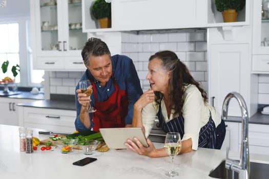 Happy senior caucasian couple in modern kitchen, cooking together, using tablet. retirement lifestyle, spending time at home.