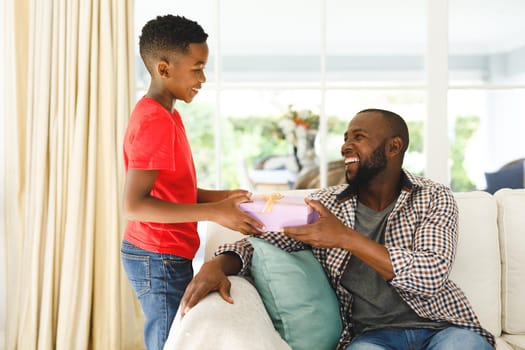 Happy african american son giving gift his father and smiling in living room. family spending time at home, father son relationship.