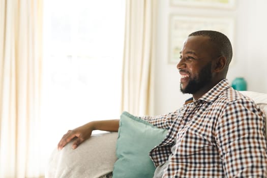 African american man smiling and sitting on couch in living room. spending time alone at home.