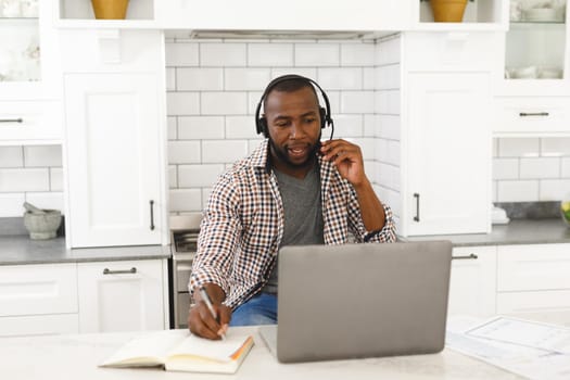 African american man sitting in kitchen making video call using laptop and headset. remote working from home with technology.