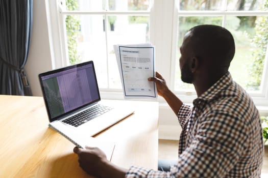 African american man sitting at table with paperwork in dining room, working remotely using laptop. flexible working from home with technology.