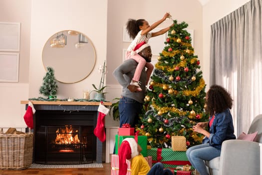 Happy african american parents and daughter decorating christmas tree. christmas, festivity and tradition at home.