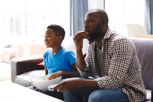 Happy african american father and son sitting on sofa, watching tv. family spending time together at home.