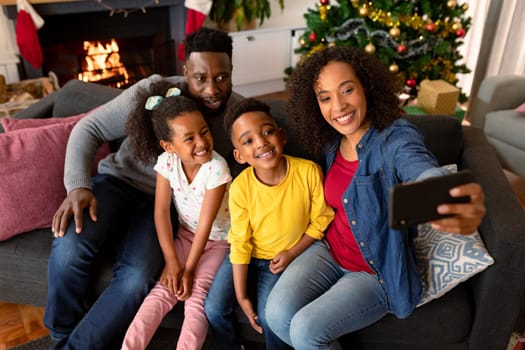 Happy african american family sitting on sofa and taking selfie, christmas decorations in background. family christmas time and festivity together at home.