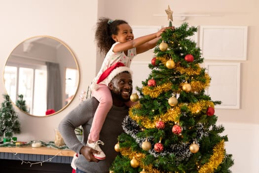 Happy african american father wearing santa hat and daughter decorating christmas tree. christmas, festivity and tradition at home.