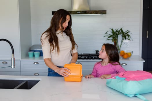 Happy caucasian mother and daughter preparing lunchbox to school in kitchen. family time, having fun together at home.