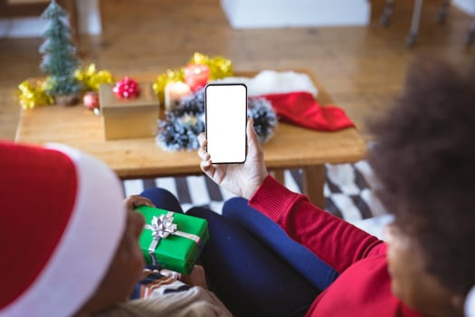 African american adult daughter and mother on christmas smartphone video call, copyspace on screen. christmas, festivity and communication technology at home.