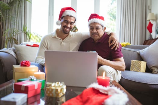 Happy biracial adult son and senior father in santa hats making laptop christmas video call. christmas, festivity and communication technology.