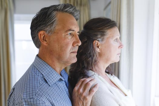 Thoughtful senior caucasian couple standing next to window, embracing. retirement lifestyle, spending time at home.