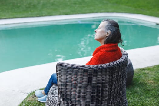 Thoughtful senior caucasian woman sitting on chair by pool in garden. retirement lifestyle, spending time alone at home.