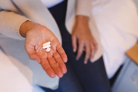 Mid section of senior caucasian woman in bedroom sitting on bed, holding pills. retirement lifestyle, illness and the aging process