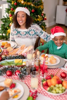 Happy african american mother and son wearing santa hats sitting at christmas table. family christmas time and festivity together at home.