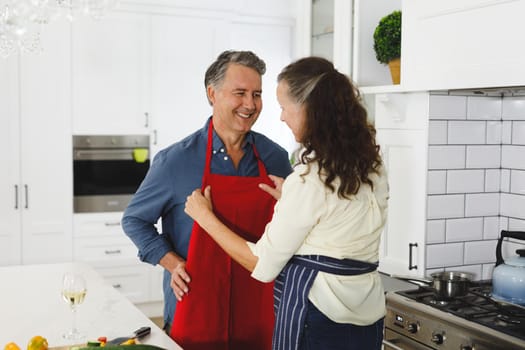 Happy senior caucasian couple wearing aprons in modern kitchen. retirement lifestyle, spending time at home.
