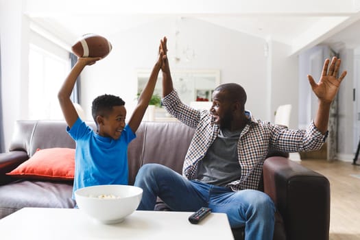 Excited african american father and son sitting on sofa, watching match on tv and cheering. family spending time together at home.