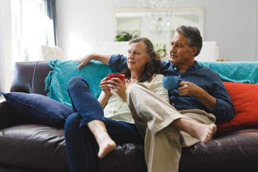 Happy senior caucasian couple in living room sitting on sofa, drinking coffee. retirement lifestyle, spending time at home.