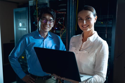 Portrait of diverse male and female engineers with laptop smiling in computer server room. database server management and maintenance concept