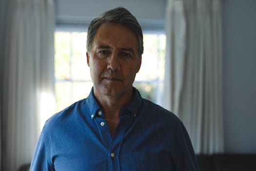 Portrait of thoughtful senior caucasian man standing next to window, looking to camera. retirement lifestyle, spending time at home.