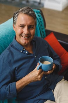 Portrait of happy senior caucasian man in living room sitting on sofa, drinking coffee. retirement lifestyle, spending time at home.