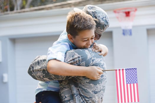 Smiling caucasian male soldier with son outside house holding american flags. soldier returning home to family.