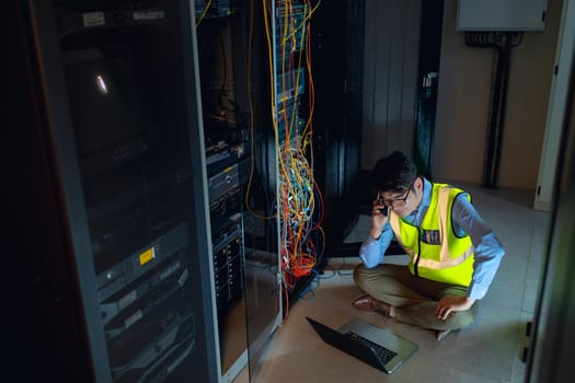 Asian male engineer with laptop talking on smartphone while inspecting in computer server room. database server management and maintenance concept