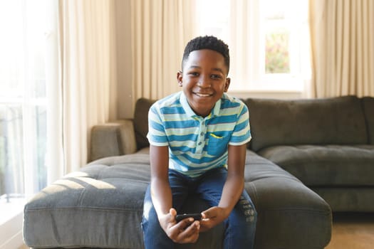 Portrait of smiling african american boy using smartphone and sitting on couch in sunny living room. spending time alone at home.