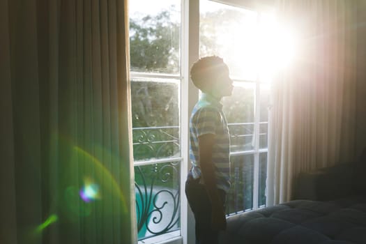 African american boy thinking and standing at window in sunny living room. spending time alone at home.