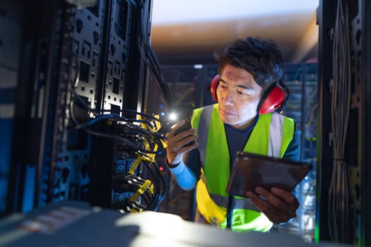 Asian male engineer using smartphone flash while inspecting the server in computer server room. database server management and maintenance concept