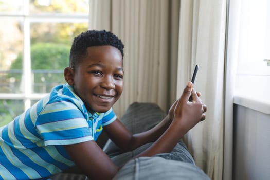 Portrait of happy african american boy kneeling on couch using smartphone in living room. spending time alone at home with technology.
