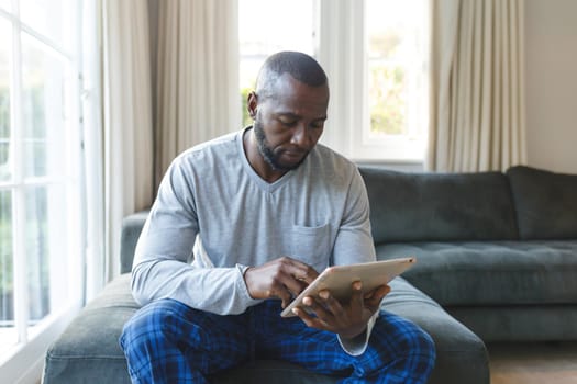 African american man using tablet sitting on couch in living room. spending time alone at home with technology.