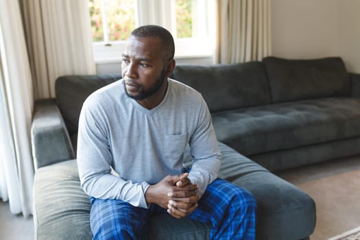 Worried, sad african american man thinking, sitting on couch looking out of window in living room. spending time alone at home.