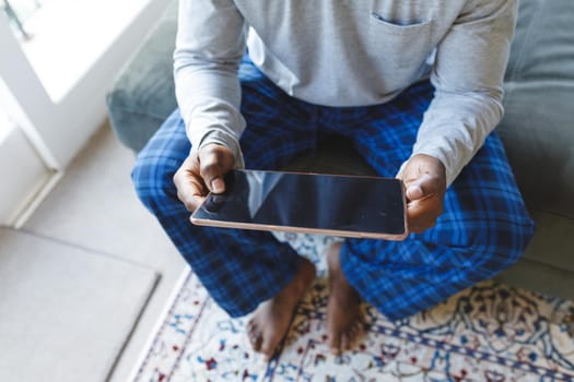 African american man using tablet and sitting on couch in living room. spending time alone at home with technology.