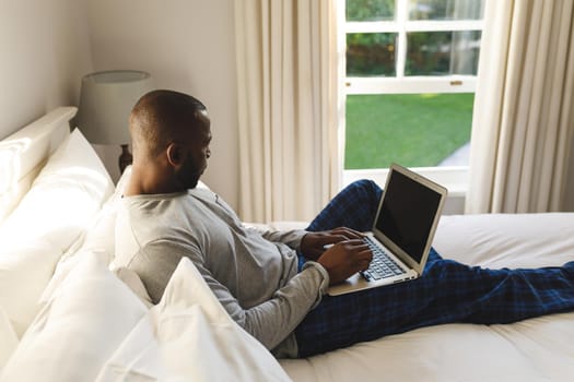 African american man using laptop and lying on bed in his bedroom. spending time alone at home with technology.