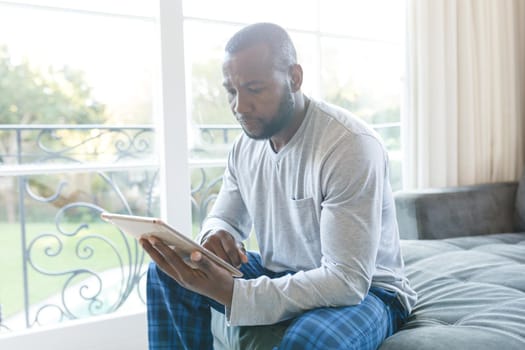 African american man using tablet and sitting on couch in living room. spending time alone at home with technology.