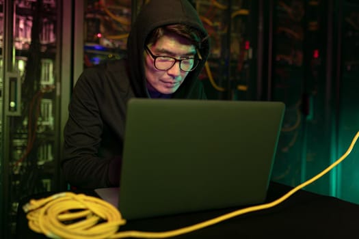 Asian male hacker using a laptop in computer server room. cyber crime and hacking concept