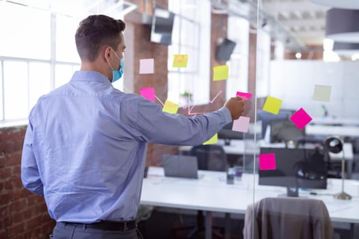 Caucasian businessman wearing face mask brainstorming, sticking memo notes on wall in office. working in business at a modern office during coronavirus covid 19 pandemic.