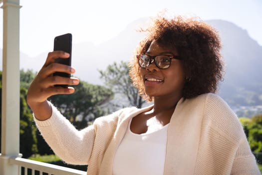 Smiling african american woman on sunny balcony of country home taking selfie with smartphone. spending free time at home.