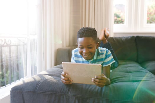 Smiling african american boy using tablet and lying on couch in living room. spending time alone with technology at home.