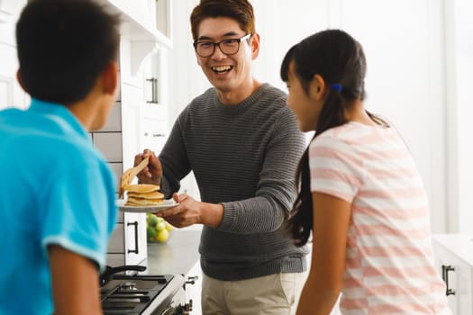Smiling asian father holding pancakes preparing breakfast with son and daughter in kitchen. family enjoying preparing meal together at home.