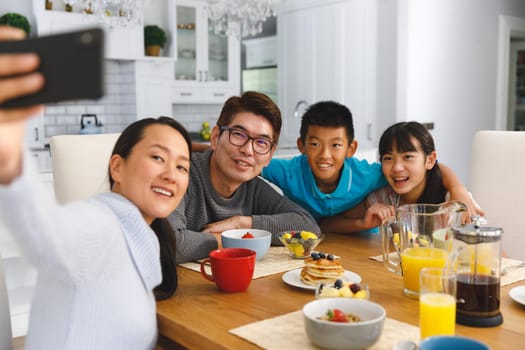 Smiling asian parents with son and daughter sitting at breakfast table taking selfie. family enjoying mealtime together at home.