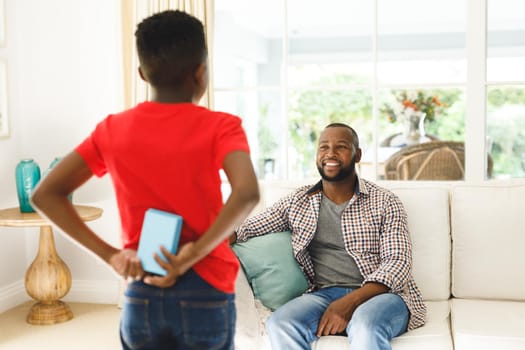 African american son giving gift his smiling father in living room. family spending time at home, father son relationship.