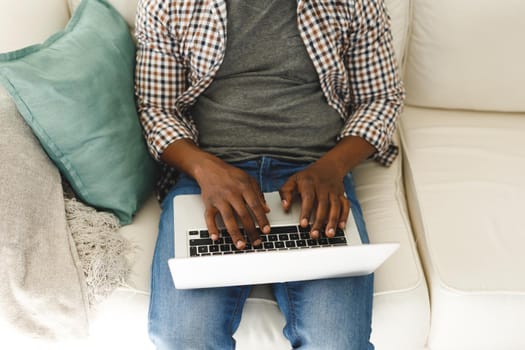 Midsection of african american man using laptop and sitting on couch in living room. spending time alone at home with technology.