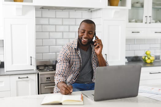 Smiling african american man sitting in kitchen working using laptop and smartphone. remote working from home with technology.