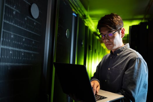 Asian male engineer using laptop in computer server room. database server management and maintenance concept