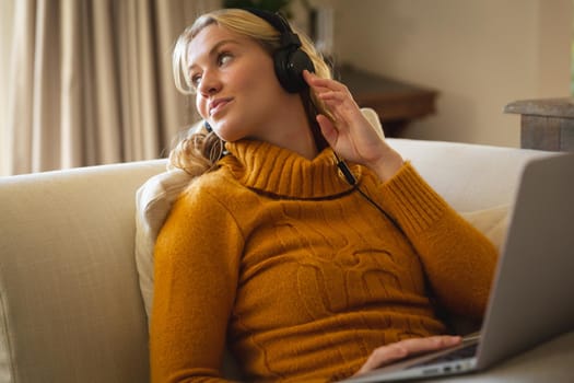 Smiling caucasian woman relaxing on couch in living room wearing headphones and using laptop. spending free time at home with technology.