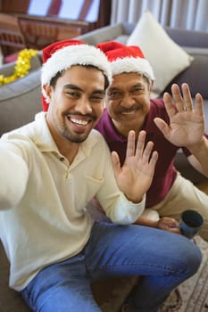 Portrait of happy biracial adult son and senior father in santa hats making christmas video call. christmas, festivity and communication technology.