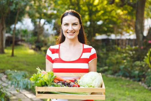 Portrait of smiling caucasian woman standing in garden holding box of fresh organic vegetables. gardening, self sufficiency and growing home produce.