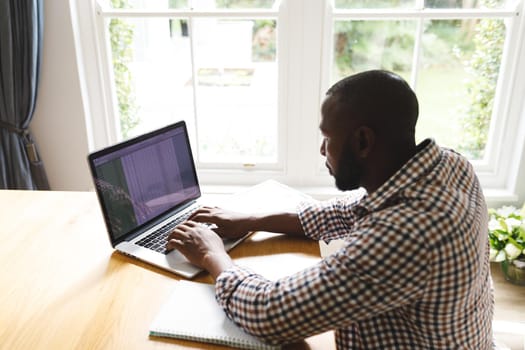 African american man sitting at table in dining room, working remotely using laptop. flexible working from home with technology.