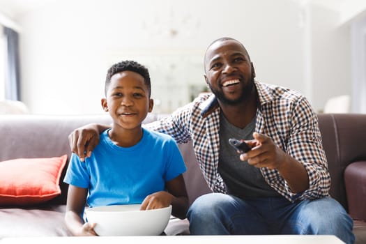 Happy african american father and son sitting on sofa, watching tv. family spending time together at home.