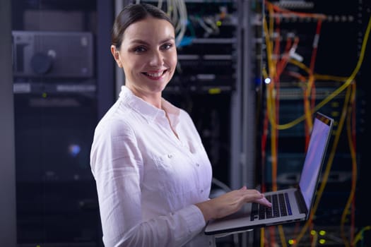 Portrait of caucasian female engineer smiling while using laptop in computer server room. database server management and maintenance concept