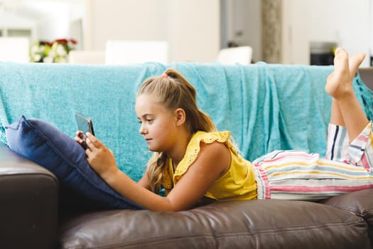 Caucasian girl lying on couch and using tablet in living room. childhood leisure time, fun and discovery at at home using technology.